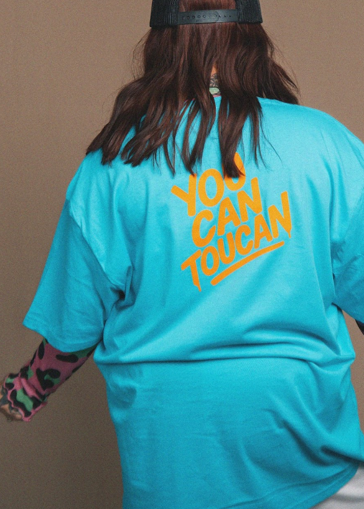 Person wearing a turquoise t-shirt with 'You Can Toucan' text on the back against a brown background
