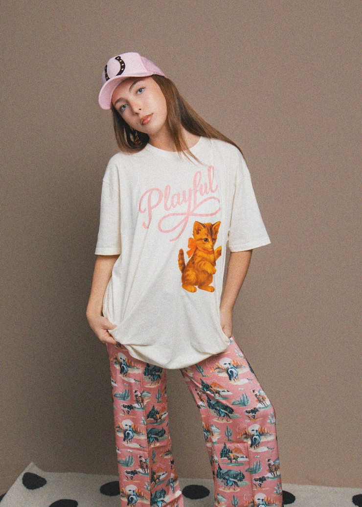 Person wearing a white t-shirt with a cat graphic and text, and patterned pants on a plain background