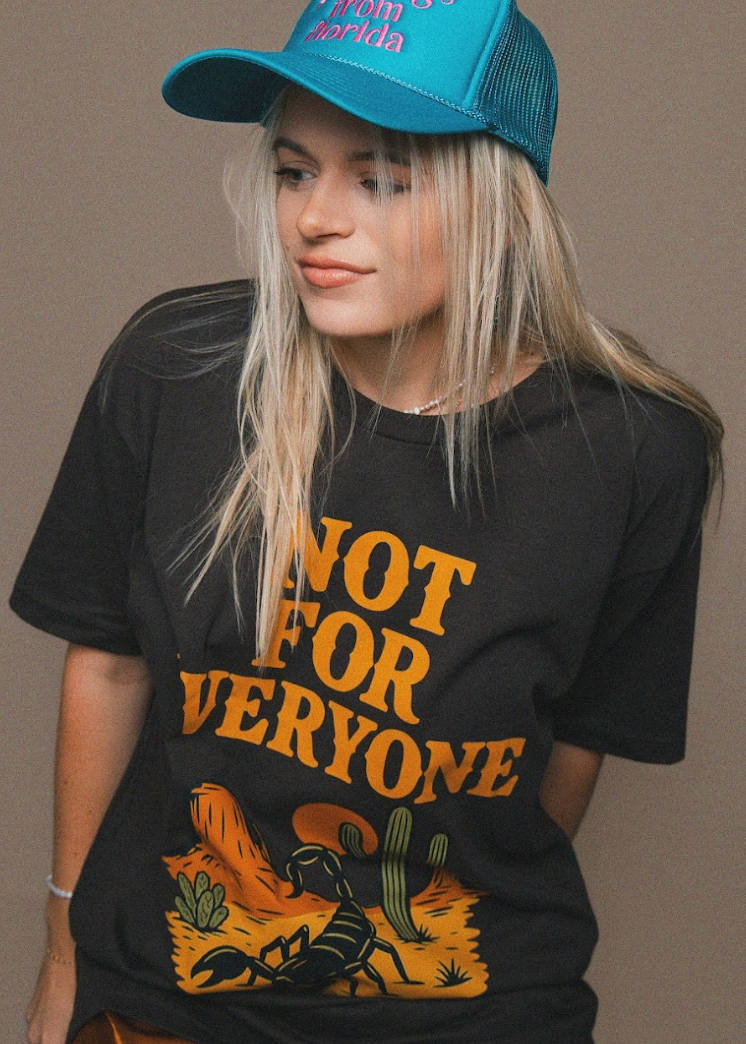 Person wearing a black t-shirt with 'Not for Everyone' graphic and a blue cap on a brown background
