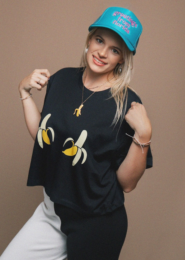 A woman posing in a black t-shirt with a banana graphic, a bright blue 'Greetings From Florida' cap, and white pants, set against a solid brown studio background.