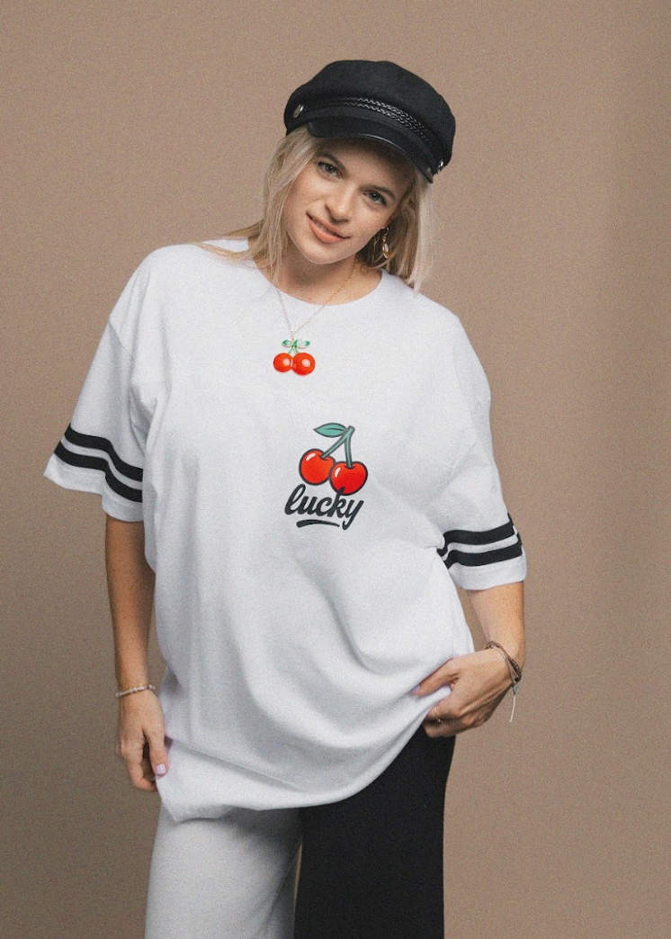 A woman modeling an oversized white t-shirt with a cherry graphic and 'Lucky Charm' text on the front, paired with a black cap, black pants, and a matching cherry necklace against a solid beige background.