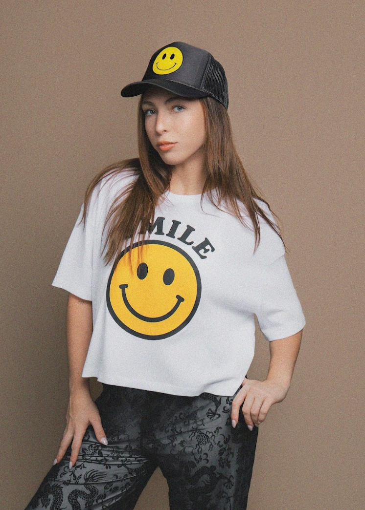 Woman wearing a white t-shirt with a smiley face design and a black cap with a yellow smiley face on a brown background