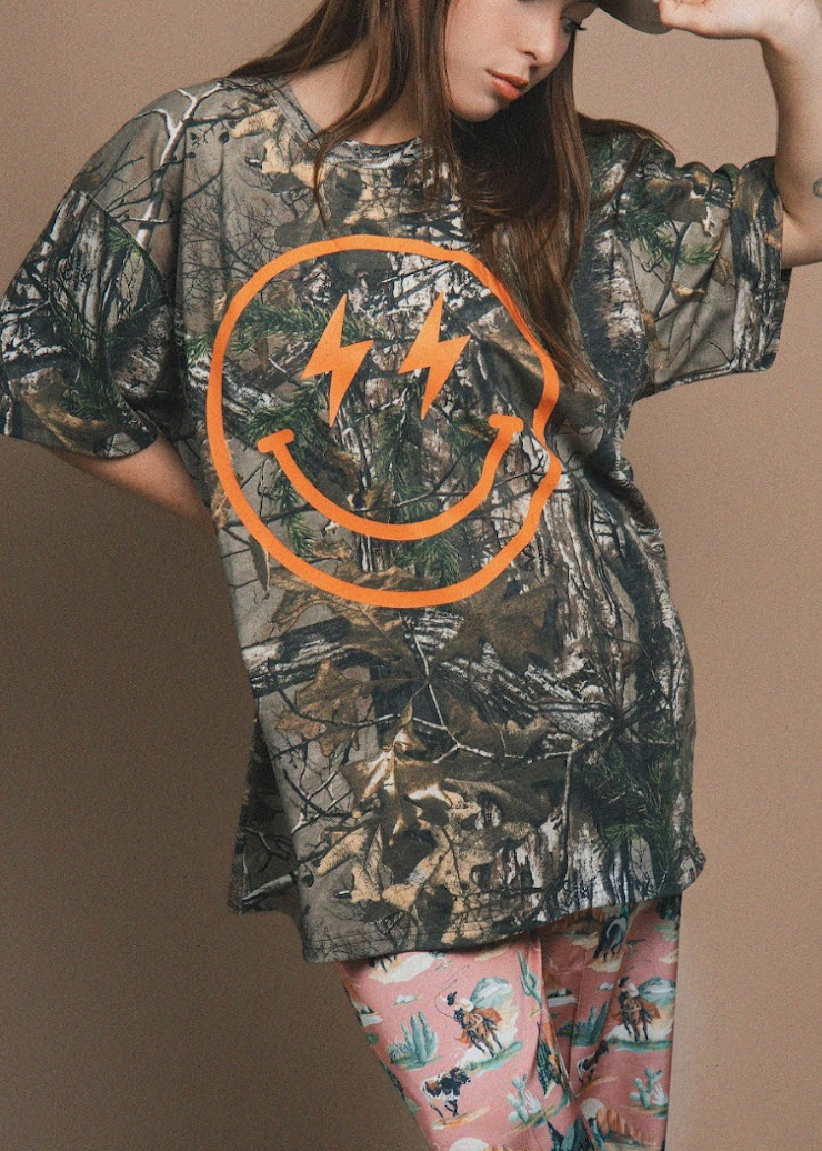 A woman wearing an oversized camouflage T-shirt with a large orange smiley face graphic containing lightning bolts, paired with pink patterned leggings, against a plain beige studio background.