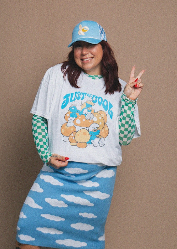 A woman modeling a Smurf-themed outfit consisting of a white graphic t-shirt with a blue and yellow cartoon print, a light blue knit skirt with white cloud patterns, and green-and-white checkered long sleeves, paired with a matching light blue cap.