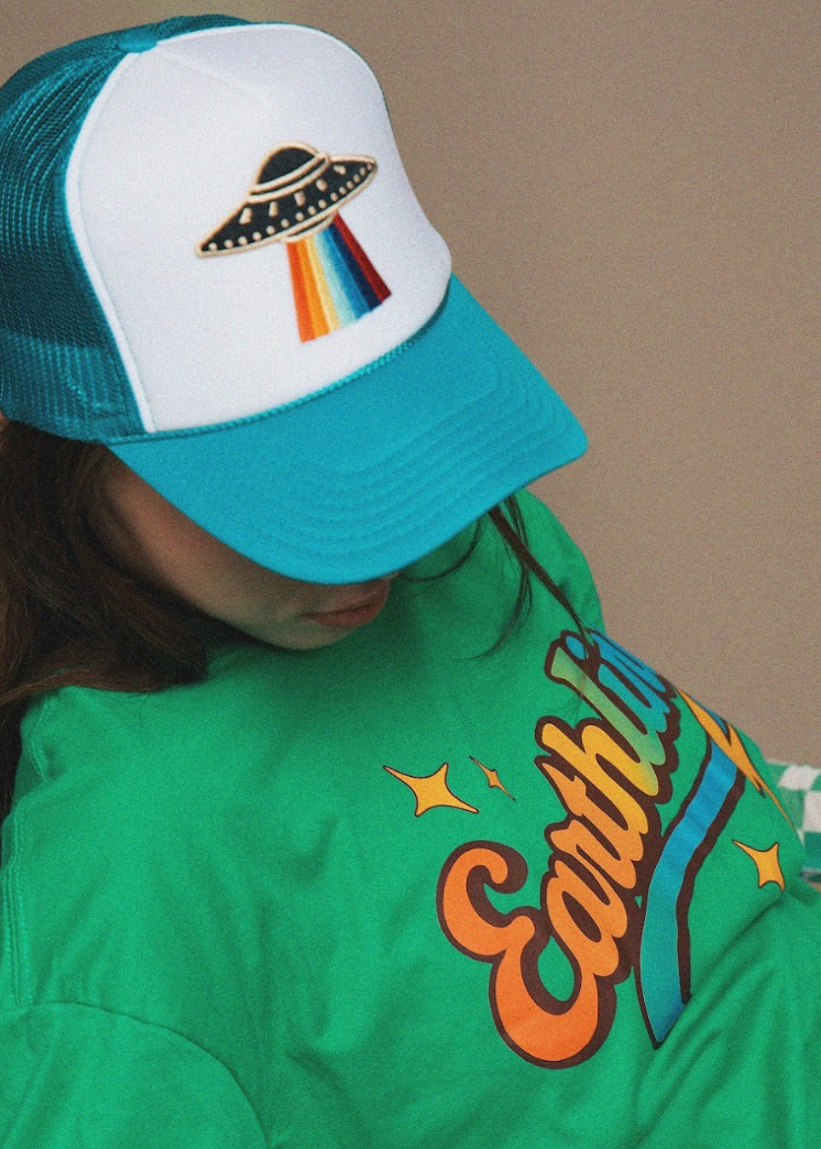 Person wearing a teal and white cap with a UFO design, standing against a beige background.