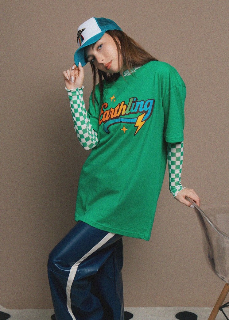 A model wearing a green graphic t-shirt over a green and white checkered long-sleeve shirt, paired with dark blue pants and a teal and white baseball cap, posing in front of a plain beige wall.