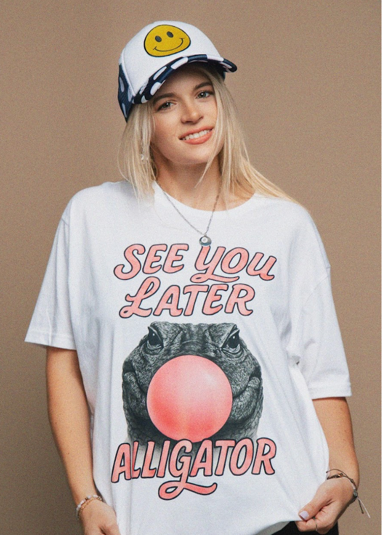Person wearing a white t-shirt with 'See You Later Alligator' graphic and a cap with a smiley face on a beige background