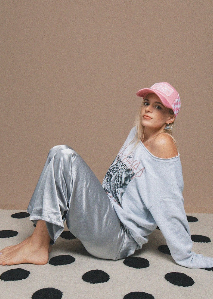 Person wearing a light grey sweatshirt and silver pants sitting on a polka dot rug with a pink cap.