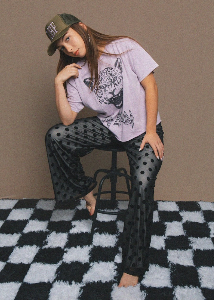 Person wearing a graphic t-shirt and polka dot pants sitting on a stool against a plain background.