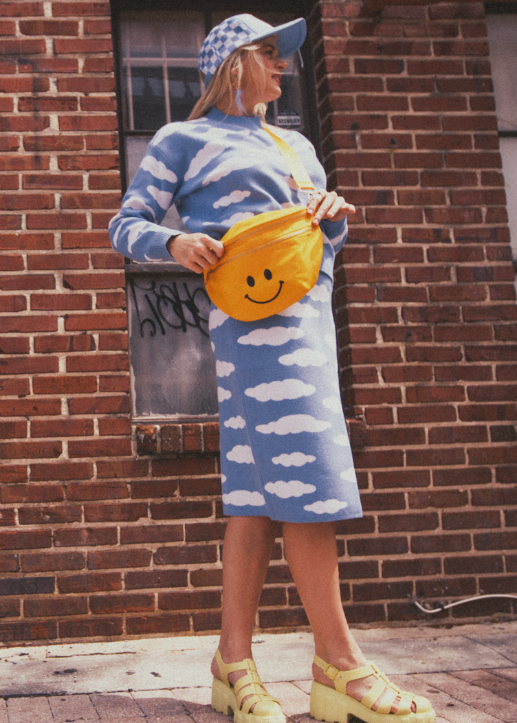 Woman in a blue set with cloud pattern holding a yellow bag with a smiley face, standing against a brick wall.