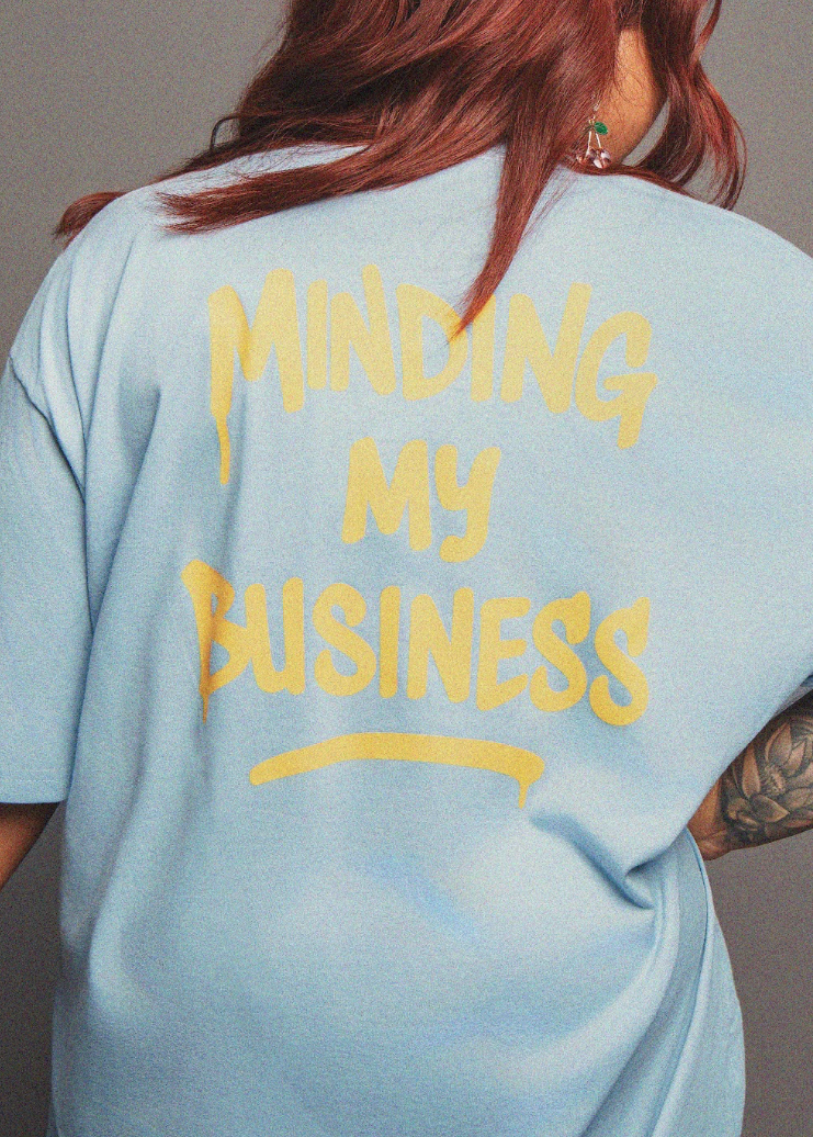 Light blue t-shirt with 'Minding My Business' text on the back, worn by a person.