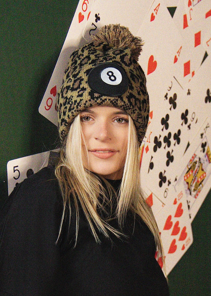 Person wearing a leopard print beanie with a number 8 ball design, standing against a playing card-themed background.