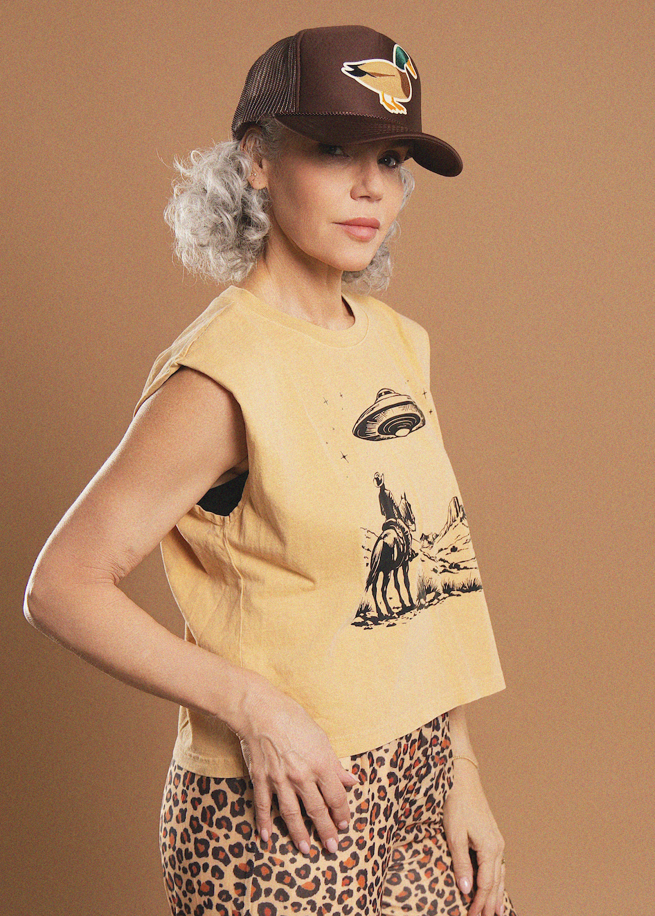 A woman modeling a cropped sleeveless t-shirt in a pale yellow color with a black graphic print of a cowboy on horseback and a UFO, paired with leopard print pants and a brown trucker hat with a coyote graphic, set against a solid tan studio background.