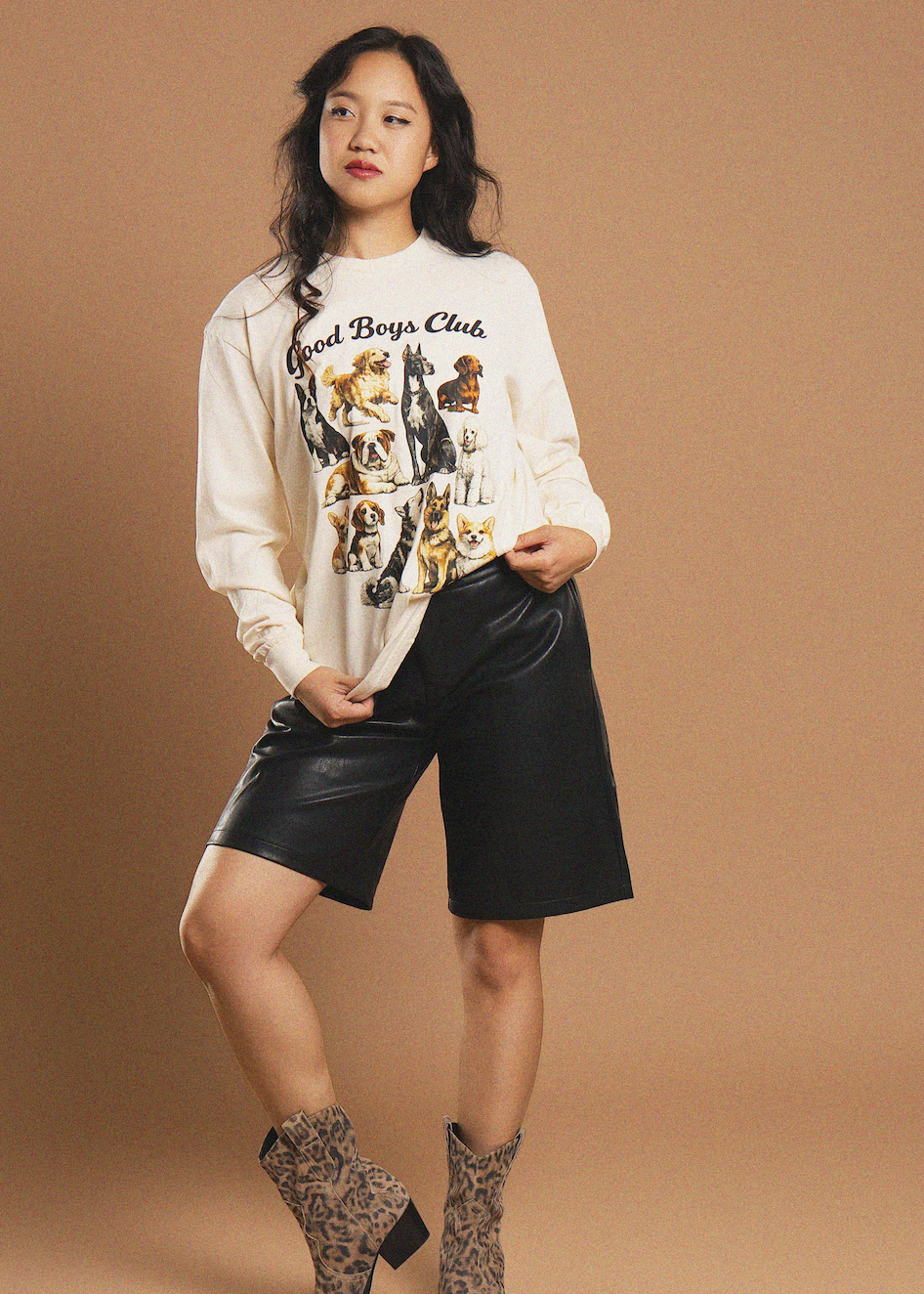 A woman models a cream-colored long-sleeve graphic sweatshirt featuring a dog print and the text 'Great Boys Club', paired with black leather shorts and leopard-print ankle boots, against a solid tan studio background.