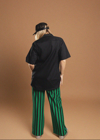 A person shown from the back wearing a black short-sleeve button-up shirt, green and black vertically striped wide-leg pants, and a black baseball cap, standing against a solid warm brown studio backdrop.