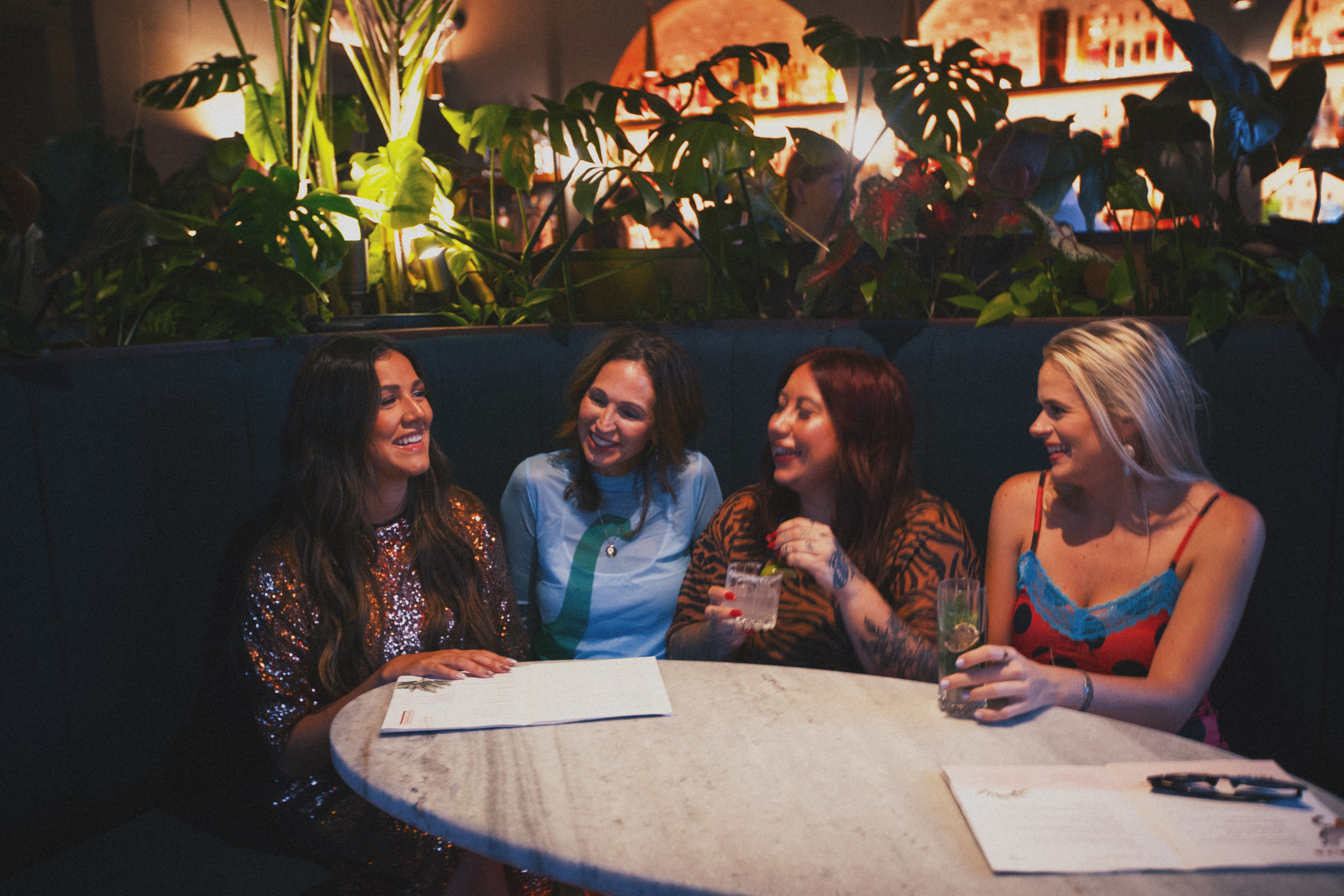 Four women sitting around a table in a dimly lit room with plants in the background