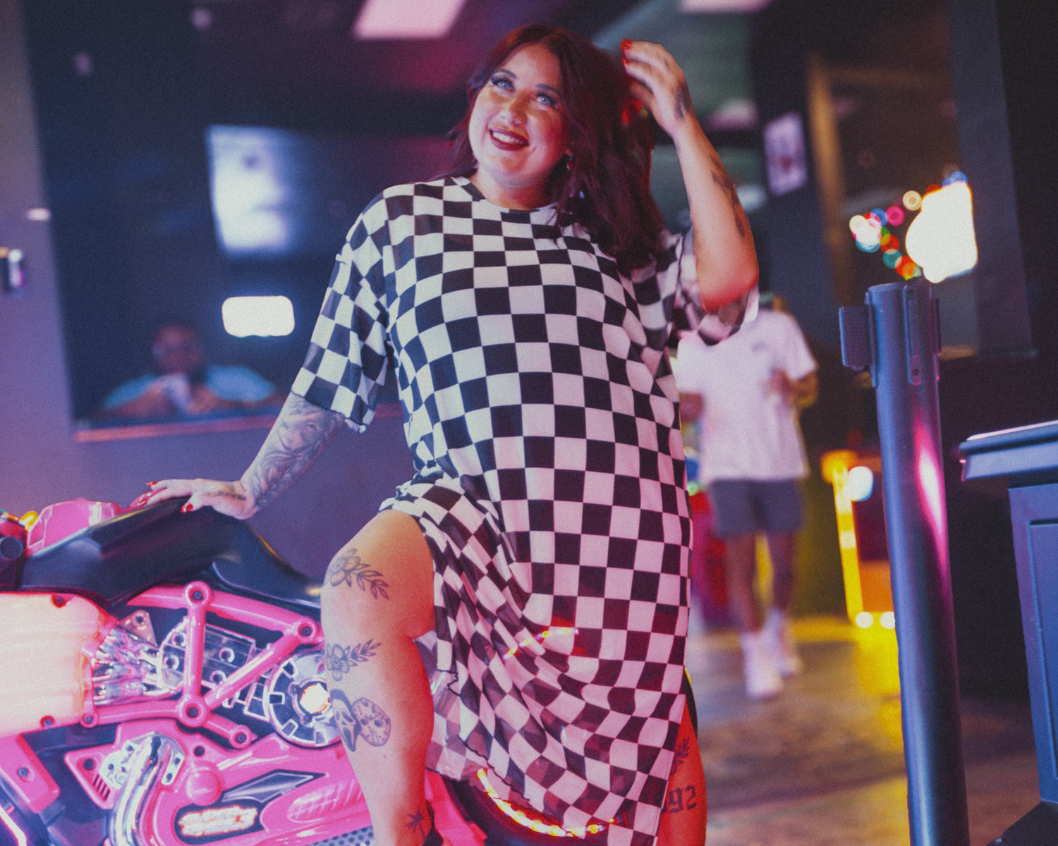 Woman in a checkered dress posing in a lively indoor setting with neon lights.