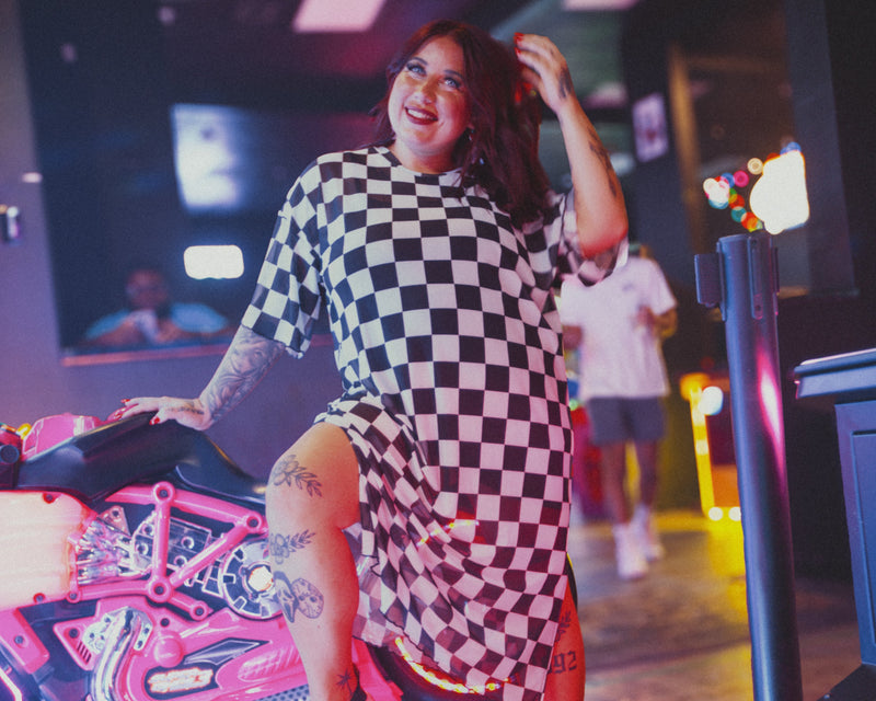 Woman in a checkered dress posing in a lively indoor setting with neon lights.