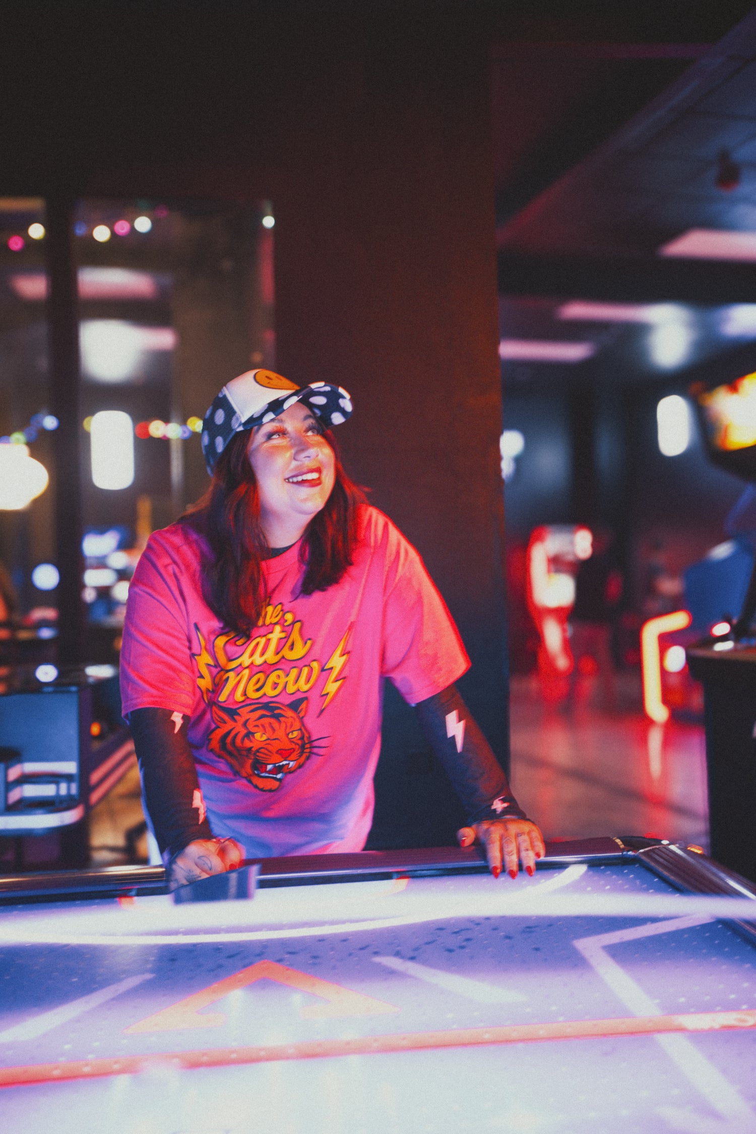 Person wearing a pink shirt with a graphic design, sitting at a table in a dimly lit room with colorful lights.