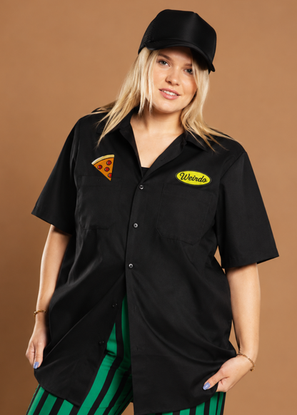 A woman modeling a black short-sleeve button-up shirt with "weirdo" patch and pizza patch and black cap against a solid brown studio background, paired with green and black striped pants