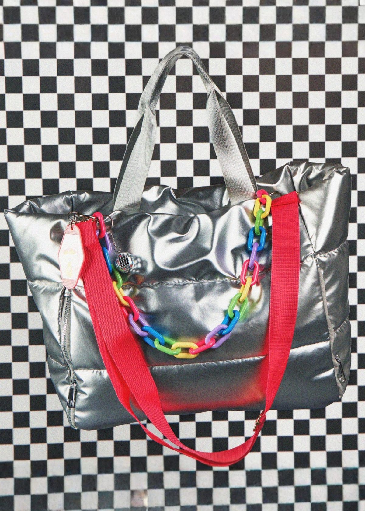 metallic silver duffle totle with hot pink strap and multi color chain strap and silver hand straps on a white and black checkered background