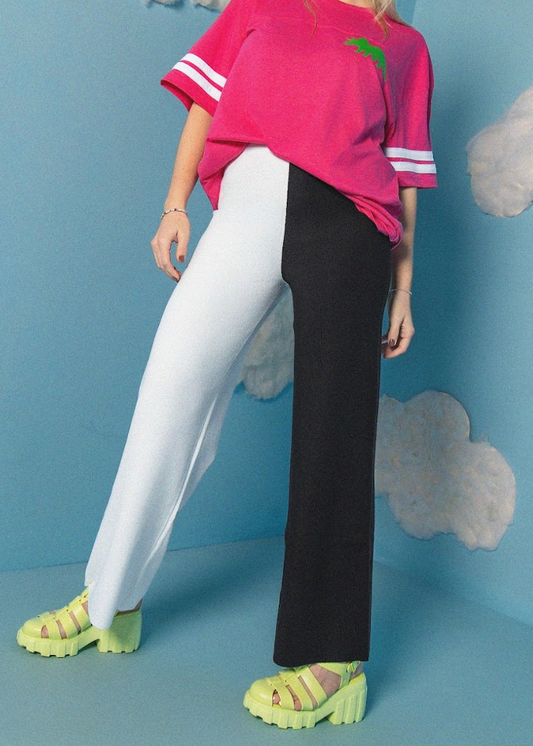 Person wearing a pink oversized shirt, white and black pants with green shoes on a blue background.
