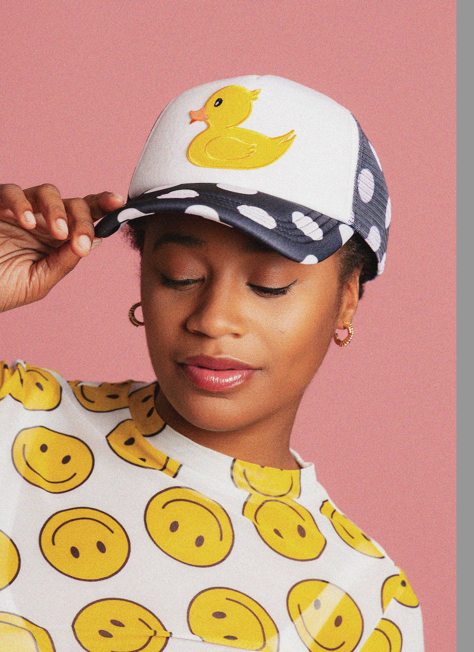 Person wearing a cap with a duck design and a shirt with smiley face patterns against a pink background