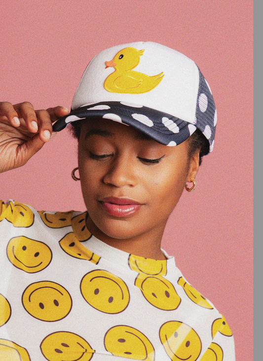 Person wearing a cap with a duck design and a shirt with smiley face patterns against a pink background