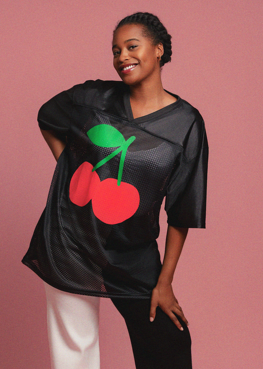 Person wearing a black mesh top with red cherries and a green stem on a pink background