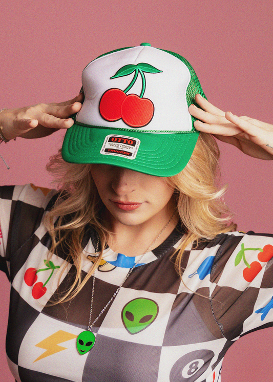 Person wearing a cap with cherry design and a patterned shirt on a pink background