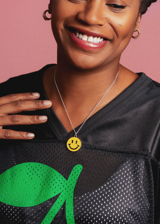 Person wearing a black sports jersey with a green logo and a yellow smiley face necklace on a pink background