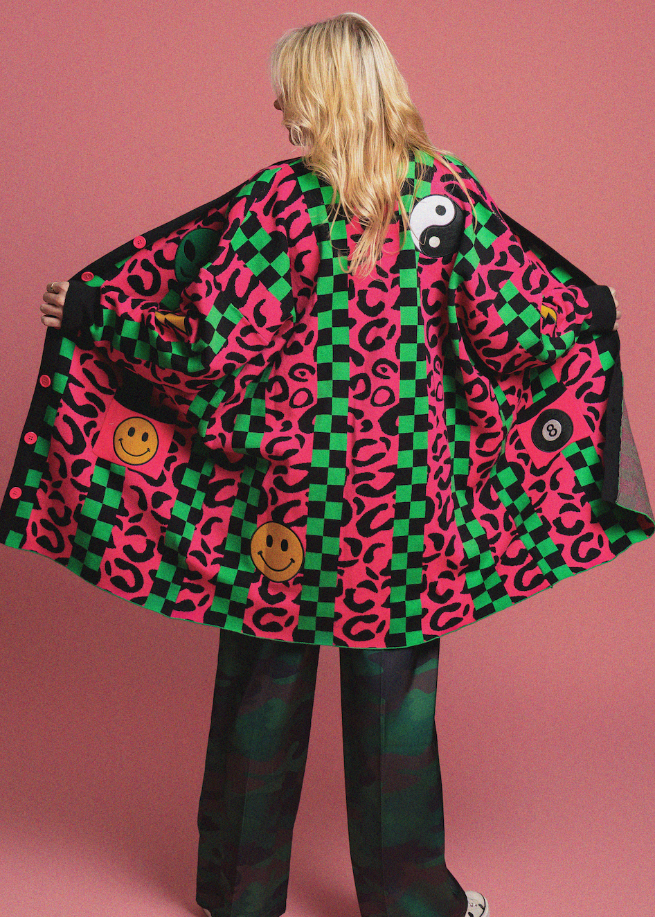 Person wearing a colorful patterned coat with smiley face patches on a pink background