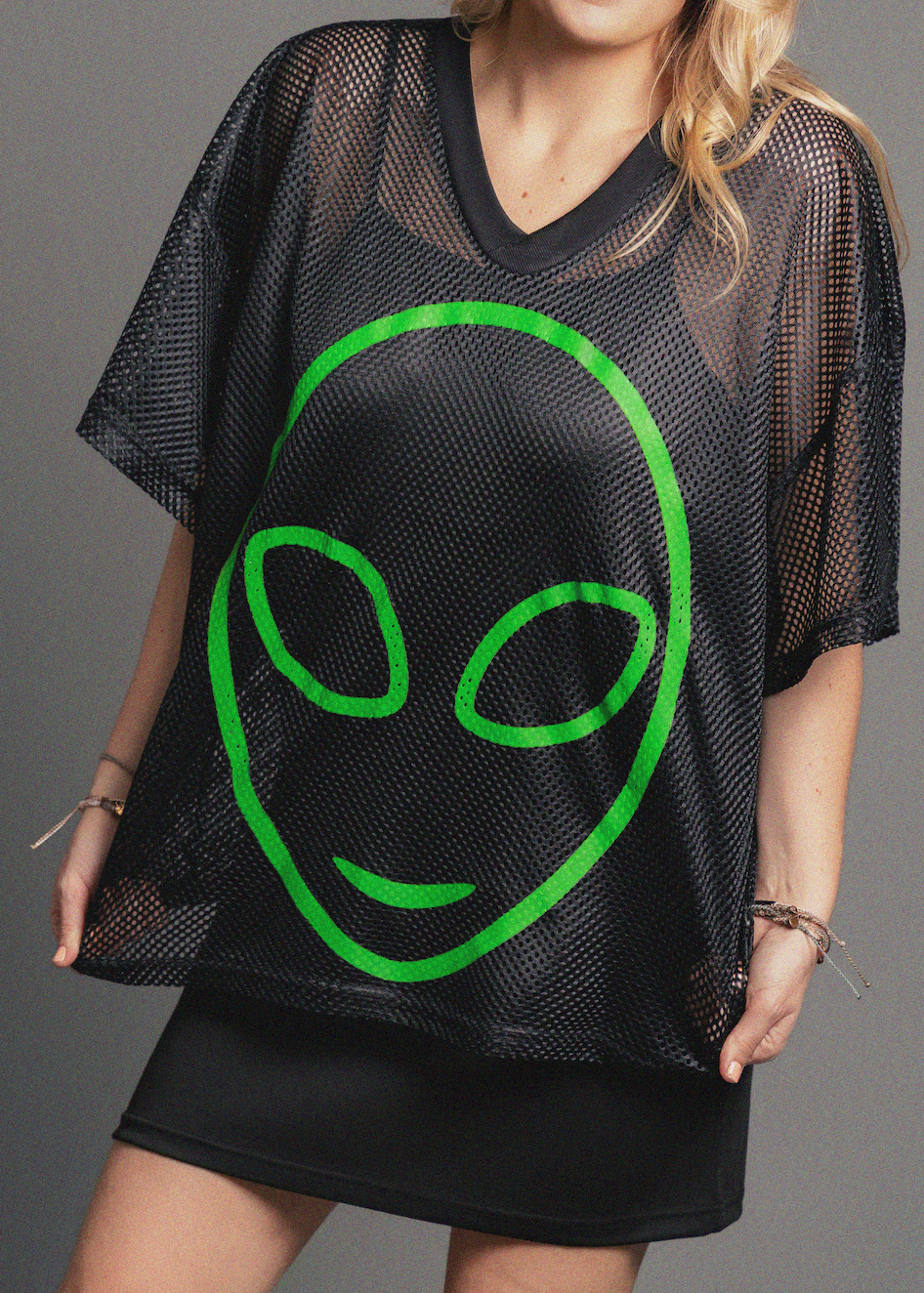 Black mesh top with green alien face design worn by a person on a gray background