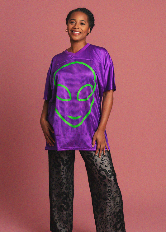 Person wearing a purple shirt with a green alien face design on a pink background