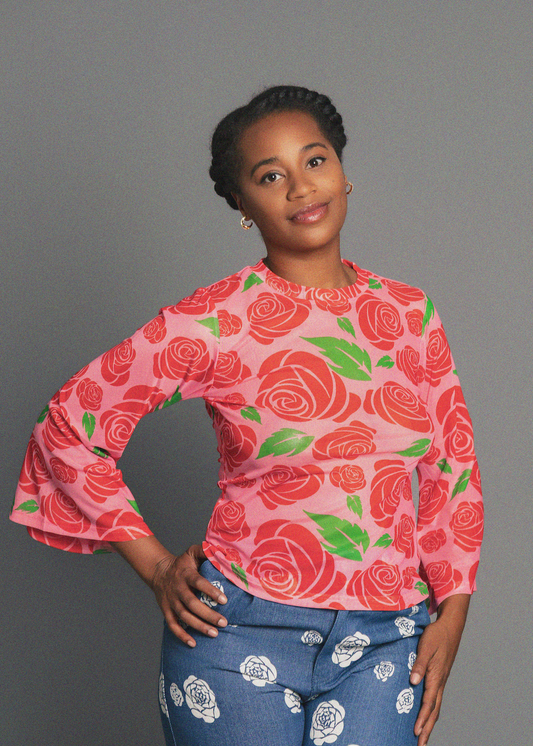 A woman posing in a pink long-sleeve top with a red rose pattern and blue jeans with white floral prints, set against a solid gray studio background.