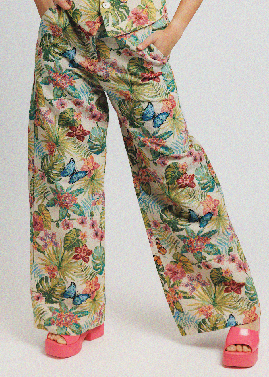 A person wearing wide-leg tropical-print pants and a matching top, paired with bright pink platform sandals, photographed against a light gray studio background.
