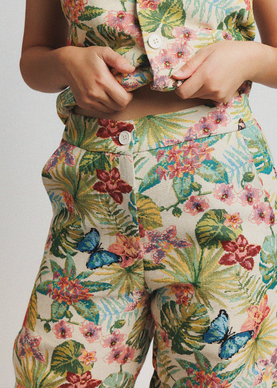 Close-up of a person wearing a matching tropical print top and pants set. The fabric features a cream base with green palm leaves, pink hibiscus flowers, red blossoms, and blue butterflies.