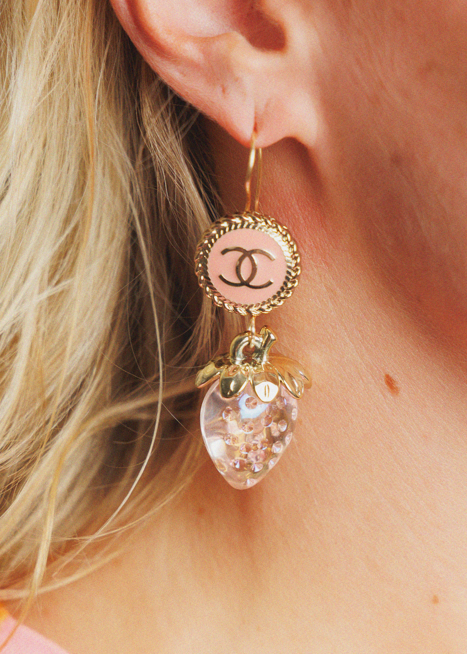 A close-up of a model's ear wearing a gold-tone dangle earring. The earring features a round pale pink charm with a black interlocking CC logo, and a dangling clear strawberry-shaped charm filled with small iridescent glitter.