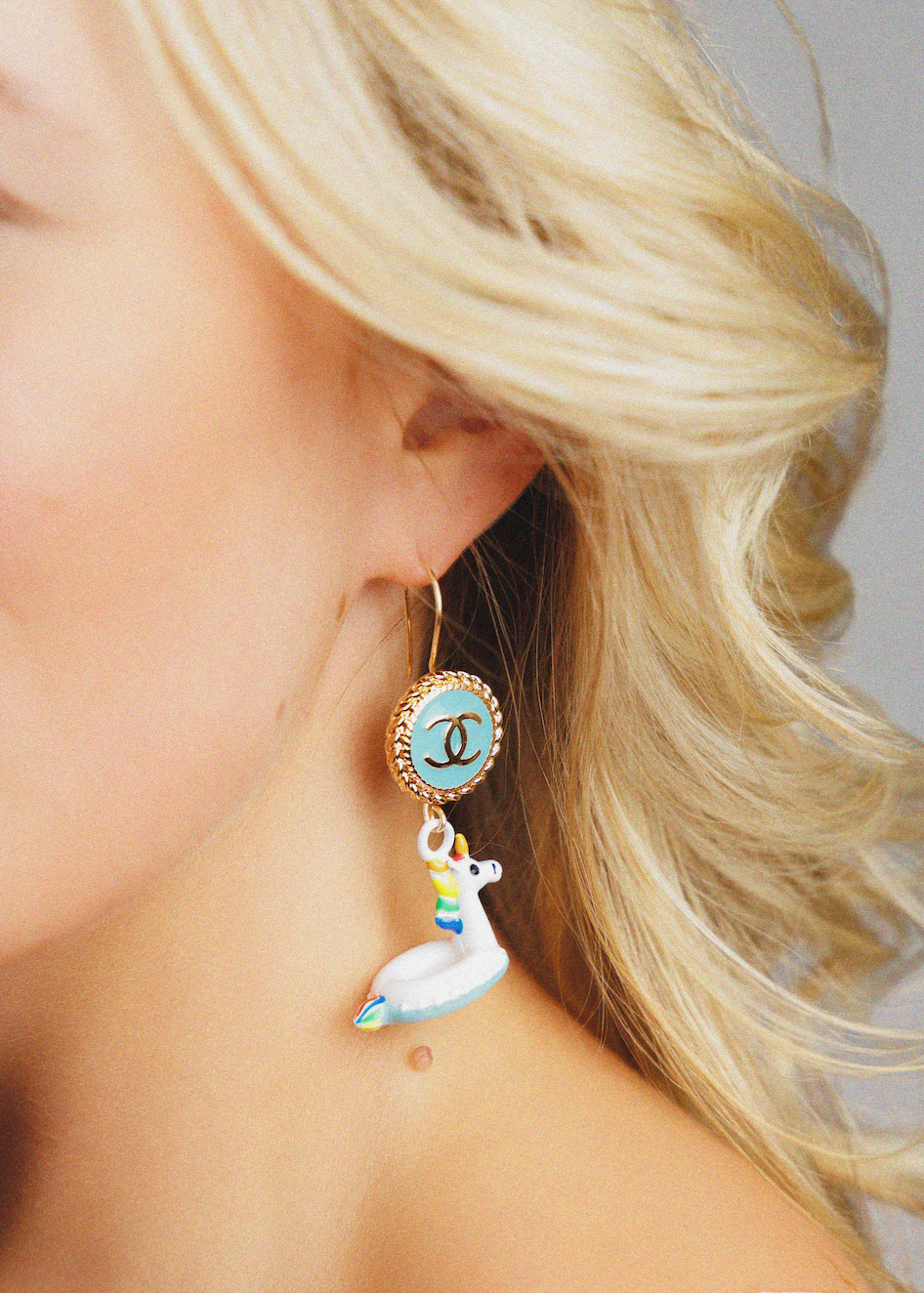 A close-up of a blonde woman wearing a dangling earring. The earring features a gold-tone top with a light blue oval insert showing a black interlocking CC logo, and a dangling white swan charm with a small rainbow accent.