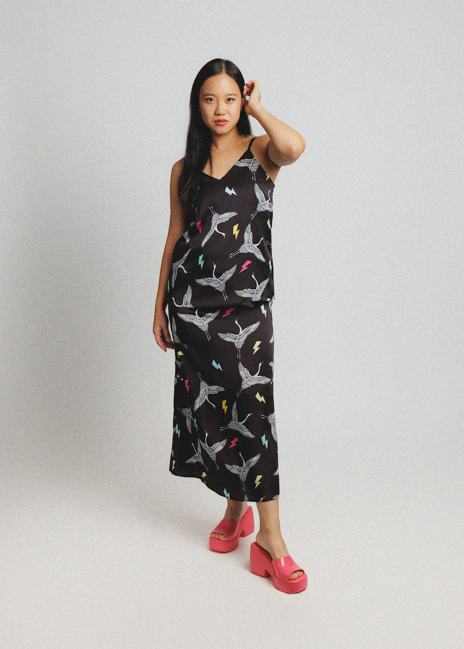A woman modeling a sleeveless black maxi dress with a white shark print and colorful accents, paired with pink platform sandals, against a light gray studio background.