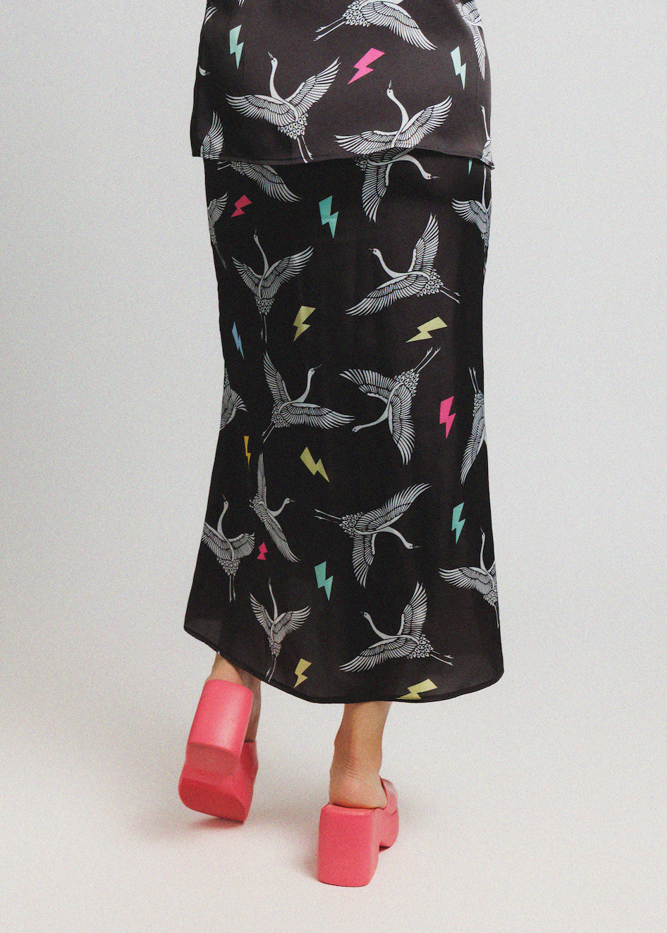 A person shown from the back wearing a black midi skirt with a white crane print and small multicolor lightning-bolt accents, paired with bright pink platform sandals, against a light gray studio background.
