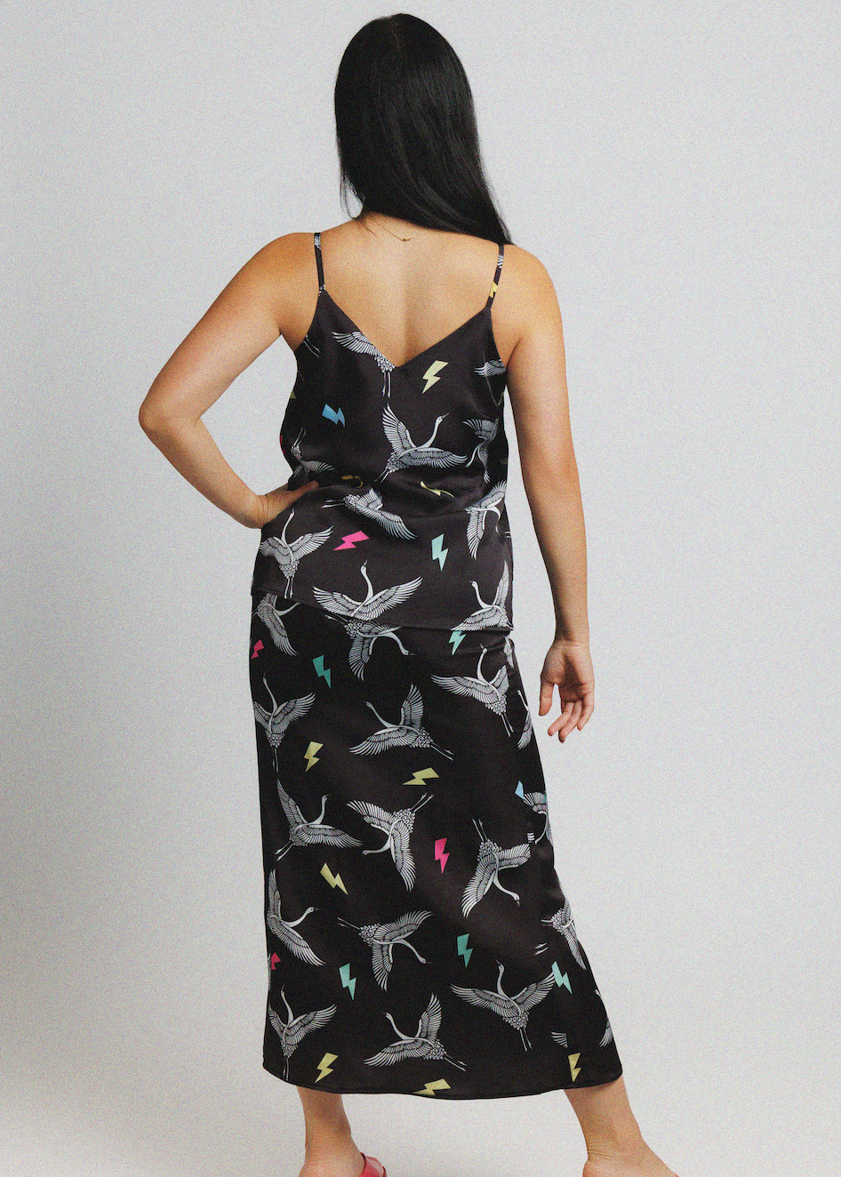 A woman shown from the back wearing a black spaghetti-strap midi slip dress with a cranes and lightning bolt pattern, set against a plain light gray studio background.