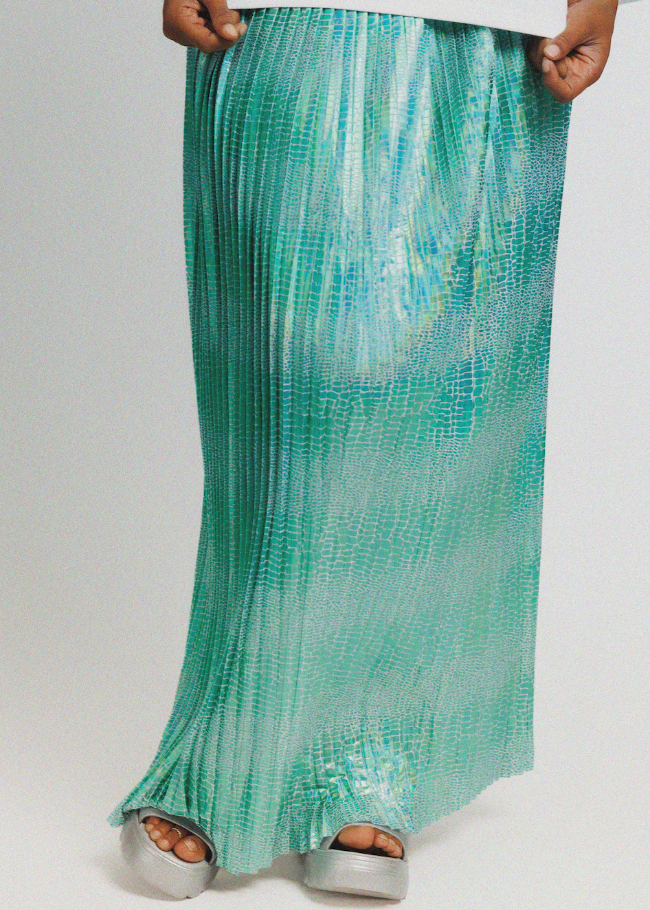 A long pleated maxi skirt in a mint green and turquoise gradient with a subtle snakeskin texture, worn by a model with silver platform sandals against a light gray background.