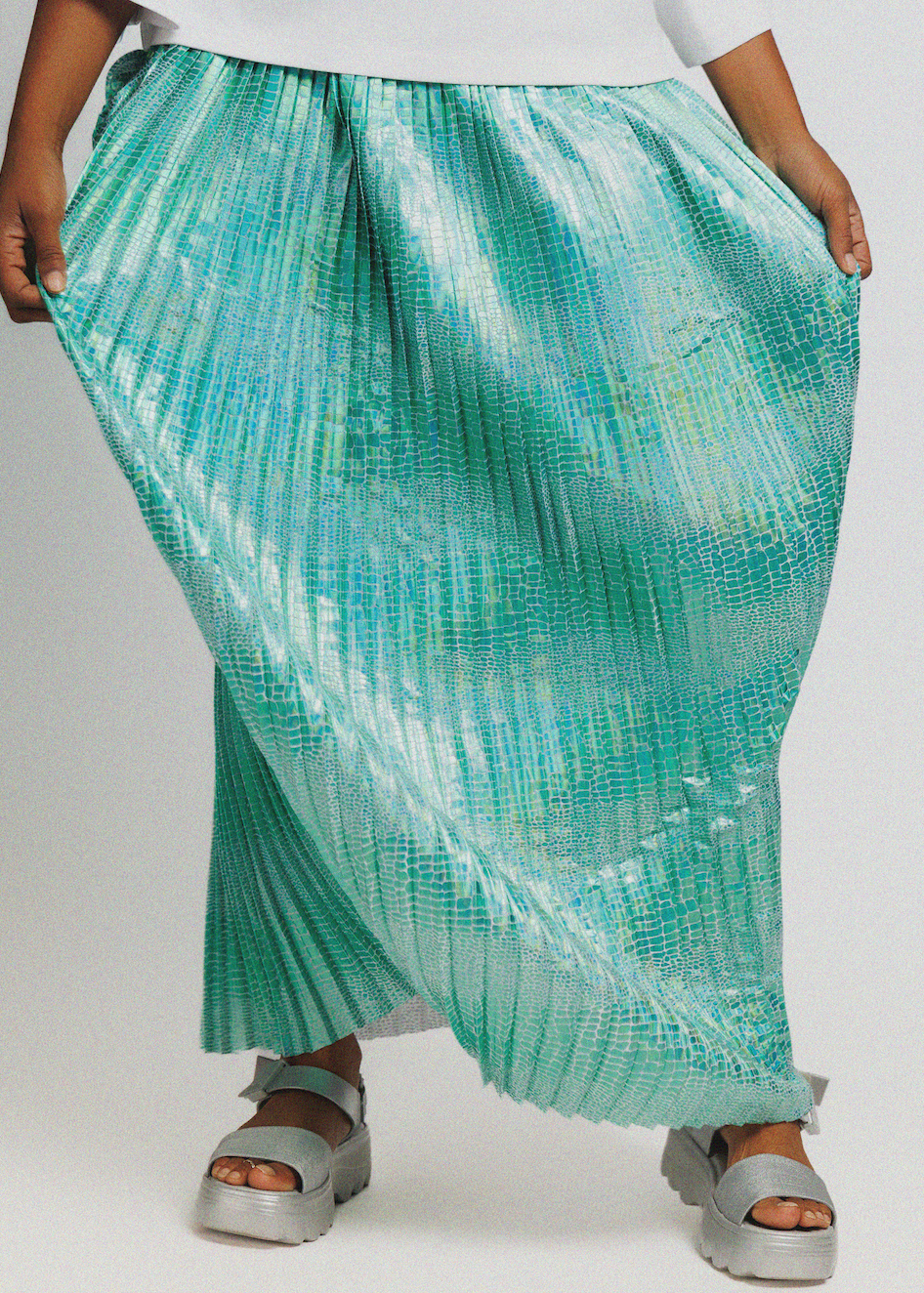 A person wearing a flowing pleated maxi skirt in a mint green and aqua color with a subtle textured pattern, paired with silver platform sandals against a light gray background.