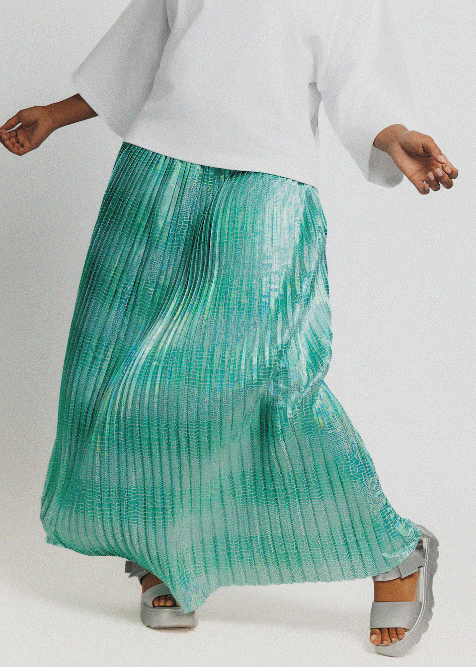 A person wearing a flowing pleated maxi skirt in a mint green to aqua gradient, paired with a white top and silver wedge sandals, captured against a light gray studio background.