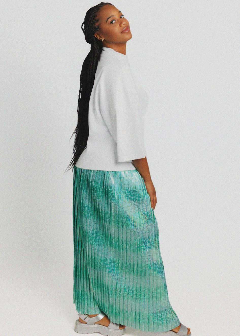 A woman shown from the back wearing a white short-sleeve top and a pleated maxi skirt in a mint green and teal tie-dye pattern, set against a plain light gray studio background.
