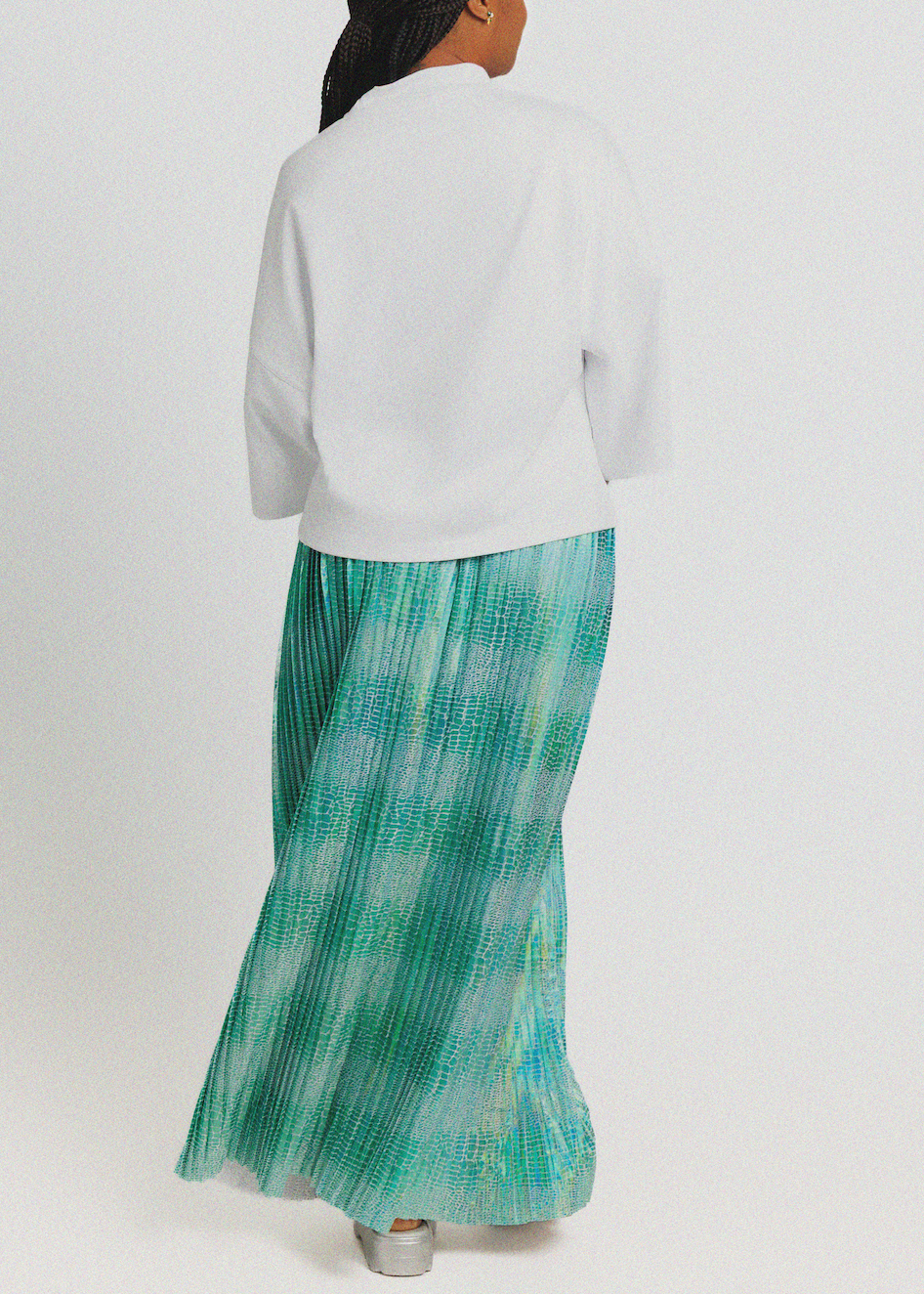 A model shown from the back wearing a white cropped jacket and a pleated maxi skirt with a green and blue tie-dye pattern, set against a plain light gray background.