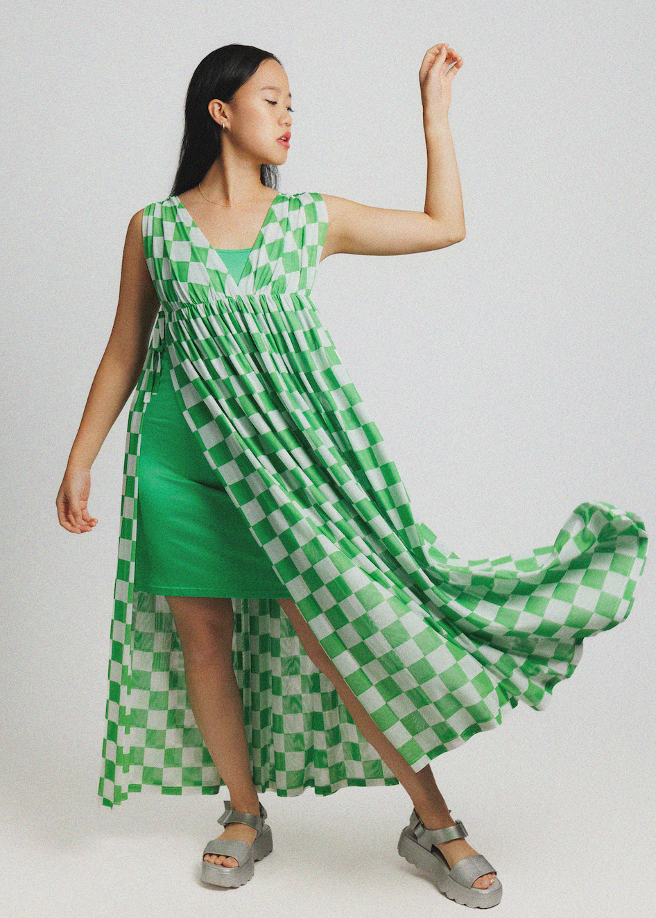 A woman modeling a sleeveless high-low dress with a green and white checkered pattern over a solid green underlayer, paired with gray platform sandals, against a light gray studio background.