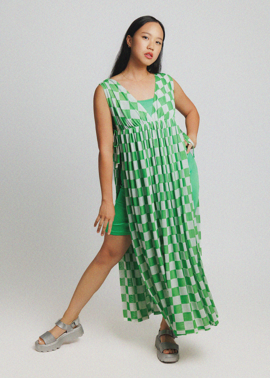 A woman modeling a sleeveless green and white checkered maxi dress with a pleated skirt and side slit, paired with gray platform sandals, against a light gray studio background.