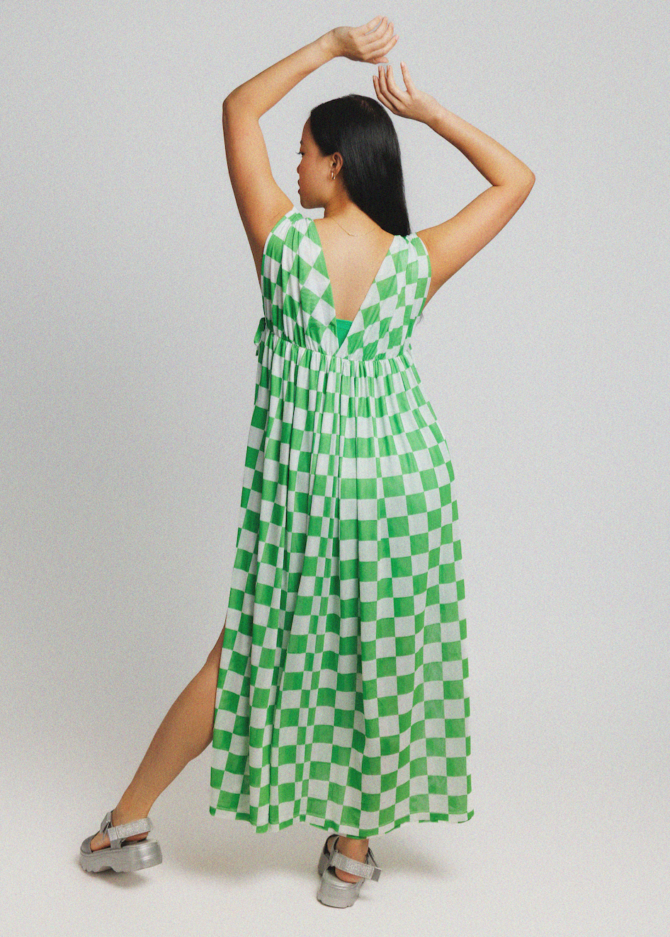 A woman modeling a green and white checkered sleeveless maxi dress from the back, posing with her arms raised against a light gray studio background.